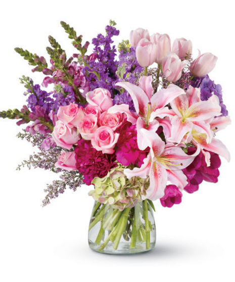 Peonies, lilies, tulips and more flowers in shades of pink and purple in a clear glass vase.
