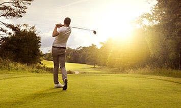 A golfer swings a club on a sunny, green golf course, surrounded by trees and bathed in warm sunlight.