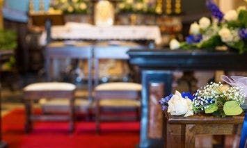 Bouquet of white and purple flowers decorates a wooden pew in a church, with two cushioned chairs and an adorned altar in the background, surrounded by additional floral arrangements and candles.
