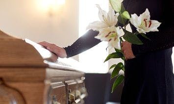 A person dressed in black holds white lilies and rests a hand on a wooden coffin in a softly lit room.