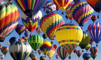 Colorful hot air balloons are floating and rising in a clear blue sky, creating a vibrant aerial display above the horizon.