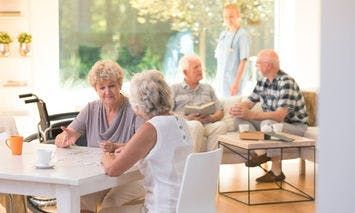 Elderly women sit at a table engaging in conversation and paperwork, while other elderly individuals and a caregiver interact in a bright, comfortable room with large windows and outdoor views.