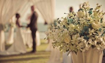 A bouquet of white flowers stands in the foreground; a couple exchanges vows in the background, framed by white drapes in a sunlit outdoor setting.